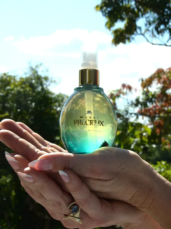 Frasco de óleo Monoi Précieux segurado delicadamente por mãos femininas com unhas feitas, em um cenário natural com árvores e céu azul ao fundo